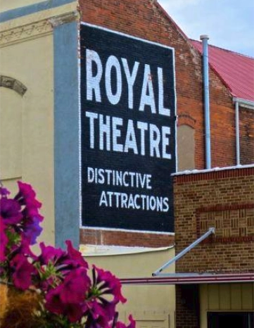 Bright purple petunias in the foreground with the historic Royal Theatre building in Macon, Missouri behind them, featuring a bold painted sign reading "Royal Theatre – Distinctive Attractions" on a red brick facade.