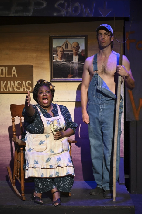 Two performers on stage during a Maples Repertory Theatre production. A woman in a floral dress and apron sits in a rocking chair, gesturing with conviction mid-scene. Beside her, a shirtless man in overalls holds a long-handled tool. A framed print of American Gothic hangs on the weathered wood backdrop behind them.
