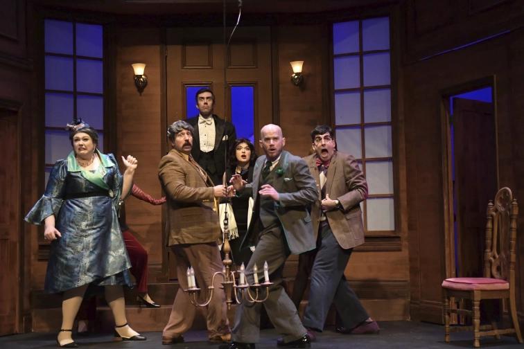 Six actors react with exaggerated shock and fear in a comedic ensemble scene on a warmly lit manor set during a live theatre production at Maples Repertory Theatre in Macon, Missouri.