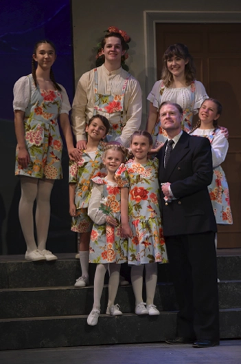 A group of children in matching floral costumes pose on stage steps with an adult actor in a suit during a kids theatre camp production at Maples Repertory Theatre in Macon, Missouri.